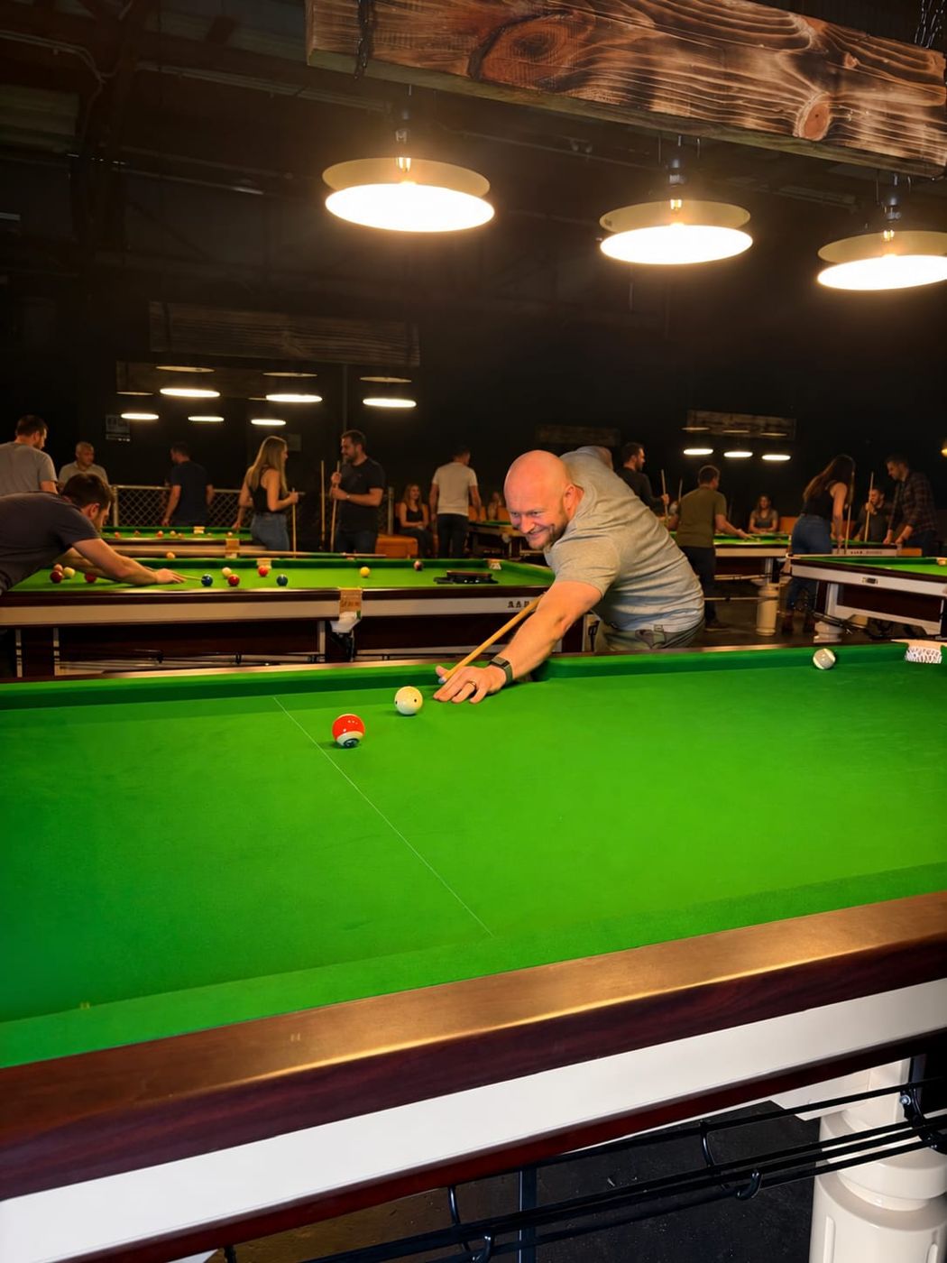Player smiling as he lines up a shot on a green pool table, with groups of people playing at tables under pendant lights behind him